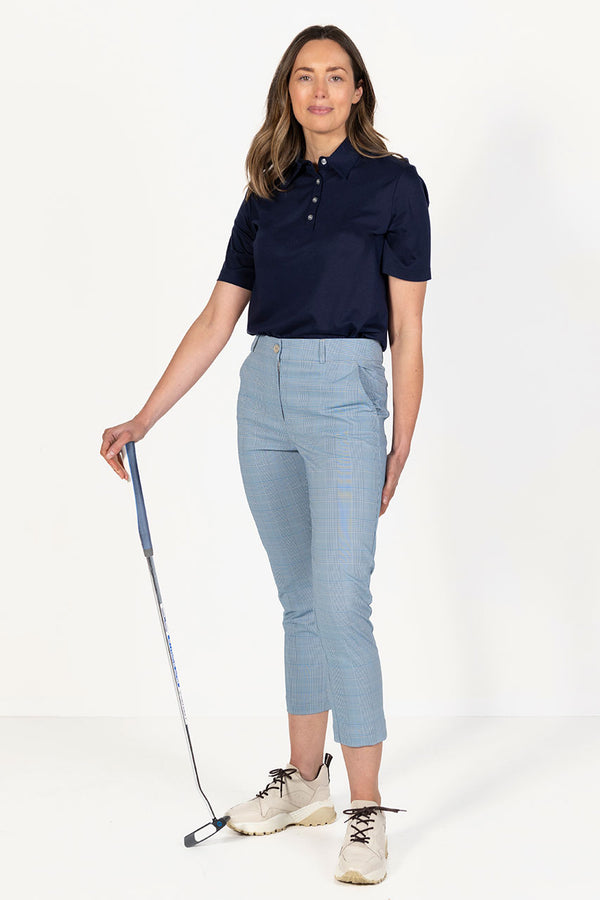 Woman wearing a navy golf polo shirt and l blue and grey check capri golf pants, holding a golf club on a white background