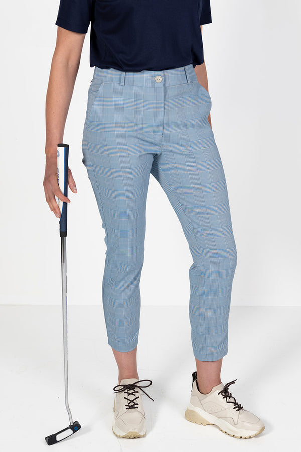 Woman wearing a navy golf polo shirt and l blue and grey check capri golf pants, holding a golf club on a white background