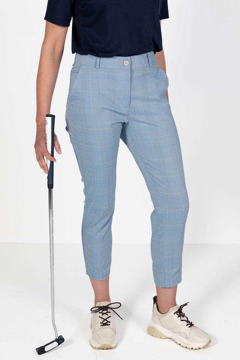 Woman wearing a navy golf polo shirt and l blue and grey check capri golf pants, holding a golf club on a white background