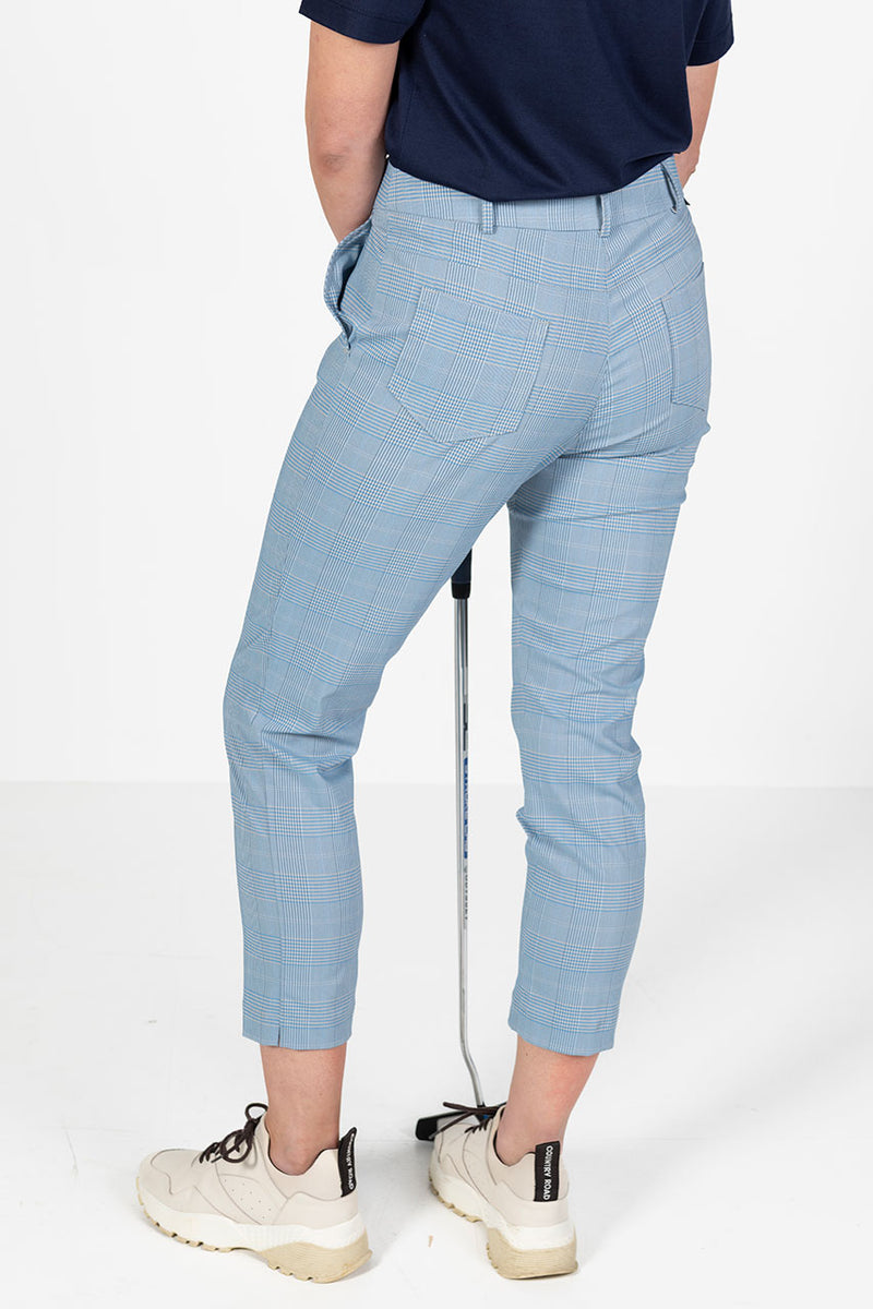 Woman wearing a navy golf polo shirt and l blue and grey check capri golf pants, holding a golf club on a white background