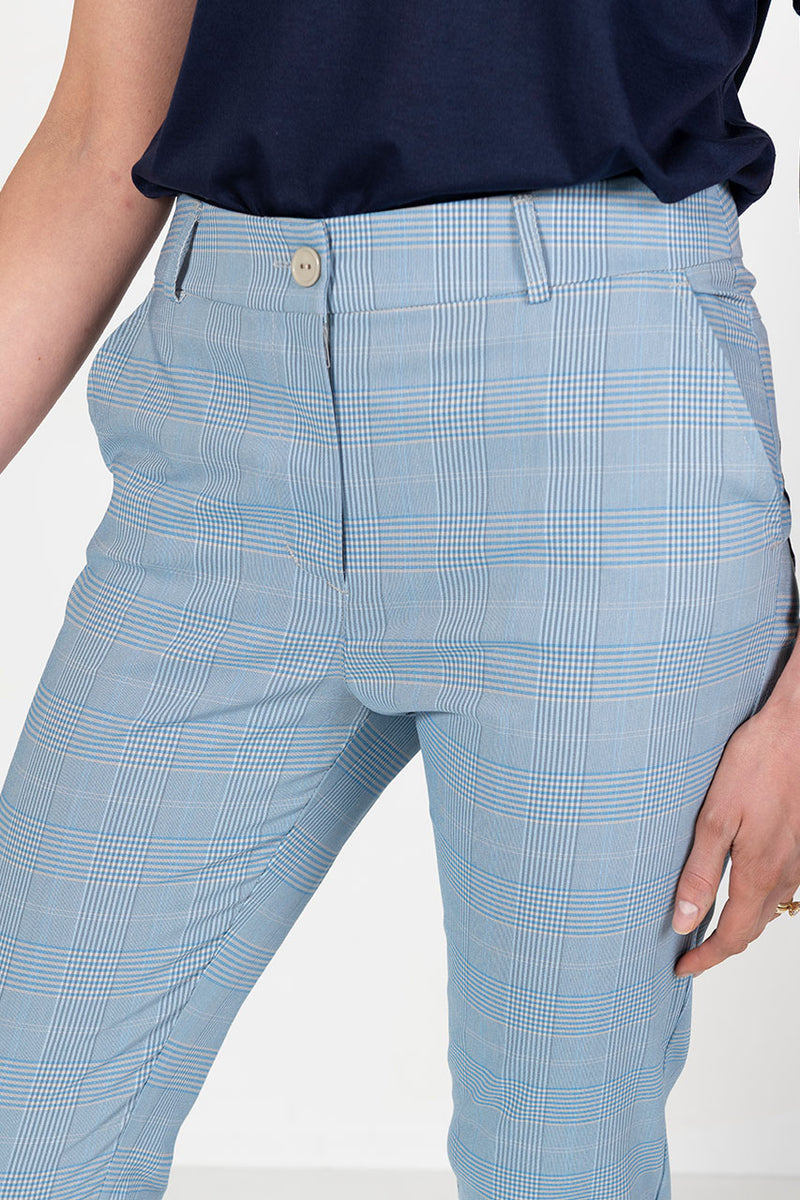 Woman wearing a navy golf polo shirt and l blue and grey check capri golf pants, holding a golf club on a white background