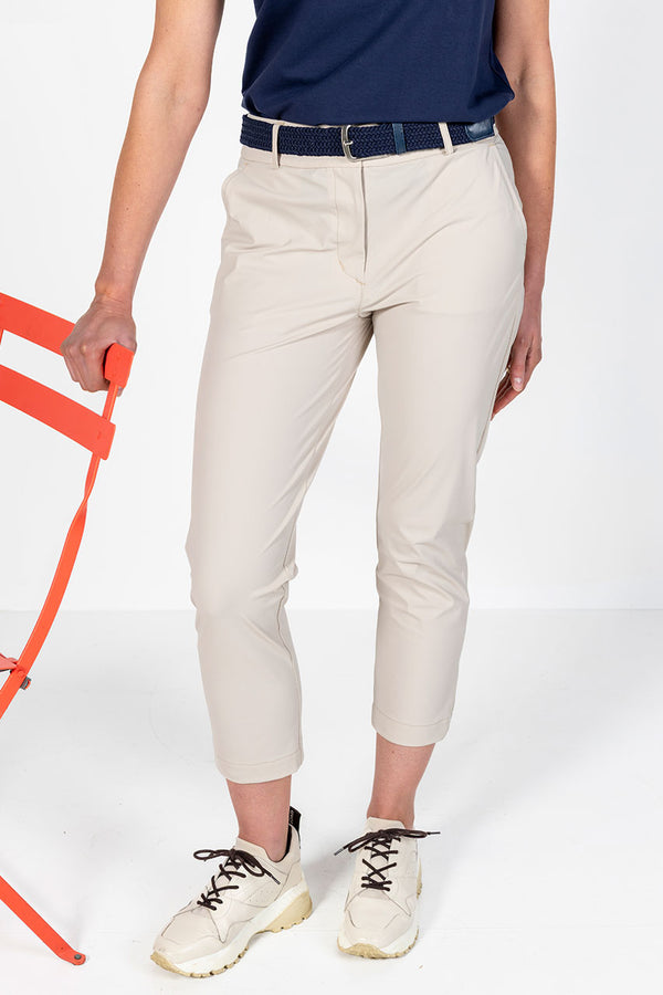 Woman in navy frill neck cap sleeve golf top and stone lightweight golf pants standing next to a orange chair on a white background